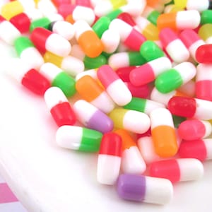 May include: A pile of colorful plastic capsules in various colors, including red, orange, yellow, green, purple, and white. The capsules are arranged in a random pattern on a white surface.