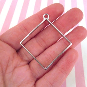 May include: A silver-colored, square-shaped pendant frame with a small loop at the top. The frame is held in a hand against a pink and white striped background. This jewelry finding is suitable for DIY projects.