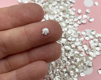 May include: A pile of silver metallic seashell sprinkles. The sprinkles are small and have a raised, textured design.