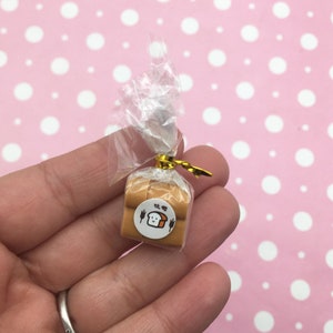 Puede incluir: Una barra de pan en miniatura envuelta en plástico transparente con una cinta dorada y una etiqueta blanca con una ilustración de pan de dibujos animados y el texto "食パン" en japonés.