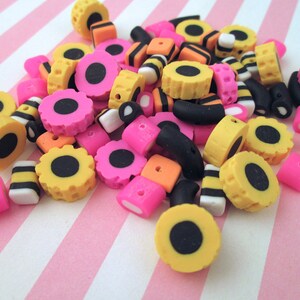 May include: A collection of colorful candy-themed polymer clay beads in black, yellow, pink, and white. The beads are shaped like licorice, jelly beans, and other candies.