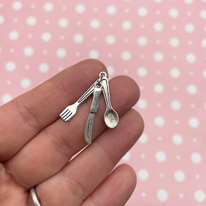May include: Silver-colored charm featuring miniature cutlery: a fork, knife, and spoon. The charm is made of a shiny metal and is designed to be a pendant or decorative element. The cutlery is detailed and realistic, suitable for jewelry or craft projects.