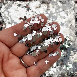 Puede incluir: Primer plano de una mano sosteniendo purpurina plateada con forma de cabezas de Mickey Mouse. La purpurina está esparcida sobre una superficie.