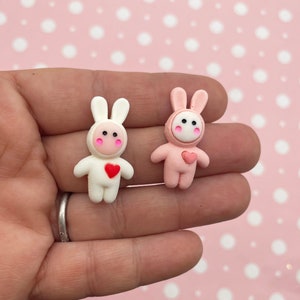 May include: Two small, handmade bunny figurines. One is white with a red heart and the other is pink with a pink heart.