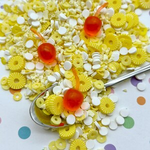 Op de afbeelding: Een close-up van een lepel gevuld met gele en witte sprinkles, met drie oranje en rode snoepkersen erop. De sprinkles liggen op een wit oppervlak met kleurrijke stippen.