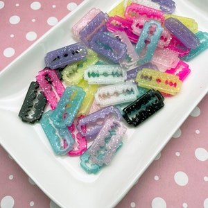 May include: A collection of colorful, glitter-covered razor blade charms in various shades of pink, purple, blue, yellow, and black. The charms are arranged in a white, rectangular dish, creating a vibrant and eye-catching display. These are ideal for crafting.