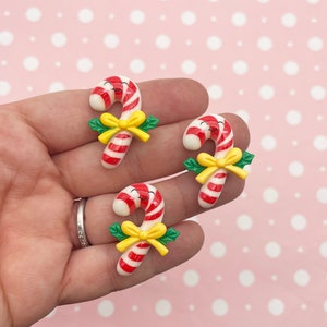 May include: Three red and white candy cane charms with yellow bows and green leaves. The candy canes have a smiling face.