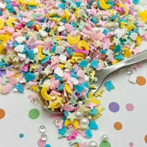 Könnte beinhalten: Eine bunte Mischung aus Streuseln mit pastellfarbenen Sternen, Monden, Wolken und anderen Formen. Die Streusel sind auf einer weißen Oberfläche mit bunten Punkten verstreut.