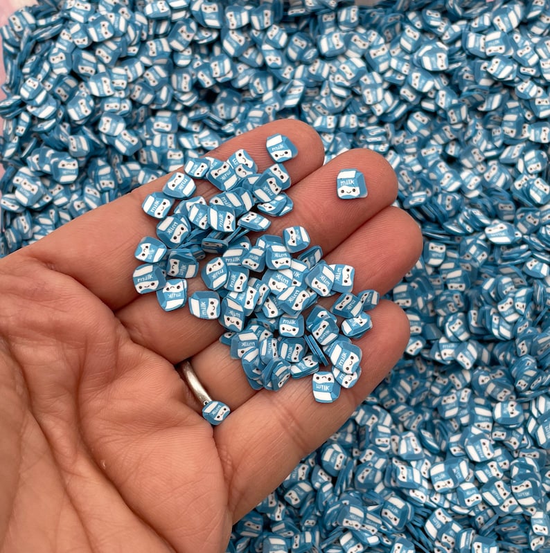 K&ouml;nnte beinhalten: Eine Hand h&auml;lt einen Haufen blauer und wei&szlig;er Milchkartonsprinkles. Die Streusel sind klein und haben ein Comic-Gesicht darauf. Der Hintergrund ist ein gro&szlig;er Haufen der gleichen Streusel.