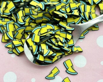 May include: A large pile of yellow and blue lightning bolt shaped resin charms with black outlines. The charms are scattered on a pink surface with white polka dots. A few charms are also on a silver spoon.