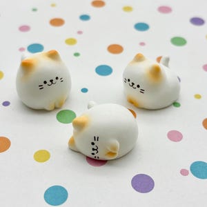 May include: Three small, white cat figurines with orange-tipped ears and simple black facial features. One cat is lying down, while the other two are upright. The background is a white surface with colorful polka dots.