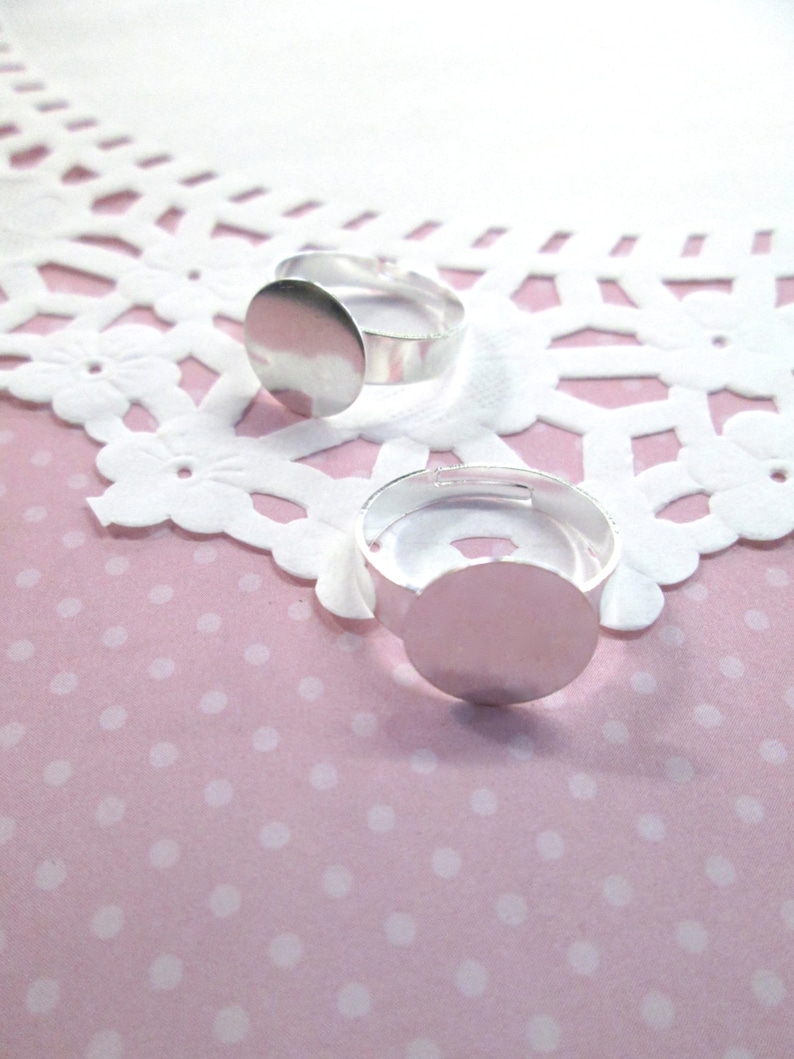 May include: Two silver ring blanks with round bezels. The bezels are smooth and shiny. The rings are on a white and pink background.