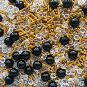 May include: A close-up of a mixture of black, gold, and clear sprinkles. The sprinkles are a variety of shapes and sizes, including small balls, long cylinders, and small squares.