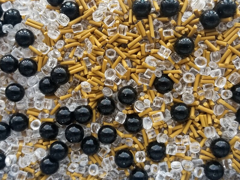 May include: A close-up of a mixture of black, gold, and clear sprinkles. The sprinkles are a variety of shapes and sizes, including small balls, long cylinders, and small squares.