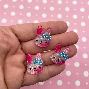 Op de afbeelding: Drie roze en witte glitterhars konijnenbedels met blauwe en witte stippen strikjes.