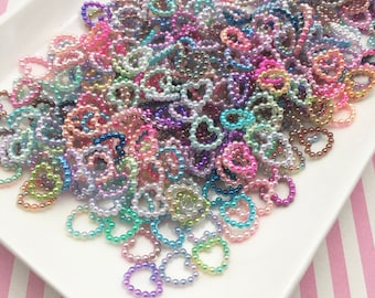 Könnte beinhalten: Ein weißer Teller gefüllt mit einer großen Menge kleiner, bunter Perlenherzringe. Die Ringe bestehen aus winzigen Perlen in verschiedenen Farben, darunter Rosa, Lila, Blau, Grün und Gold.