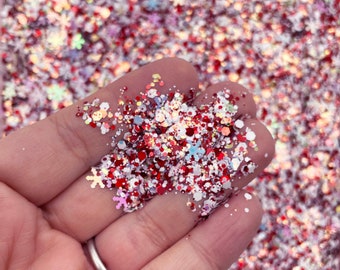 May include: A close-up of red, white, and iridescent glitter with snowflake and other shapes. The glitter is being held in a hand.