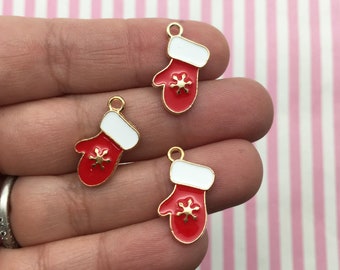 May include: Three red and white enamel mitten charms with gold-coloured trim and a snowflake design. Each charm has a small loop at the top for attaching to jewellery or other crafts. The background is a pink and white striped pattern.