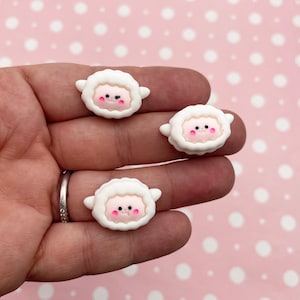 May include: Three white clay charms shaped like sheep with pink cheeks and black eyes. The sheep are approximately 1 inch in size.