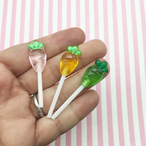 Può includere: Tre lecca-lecca in miniatura a forma di carota su bastoncini bianchi. I lecca-lecca sono trasparenti e riempiti di caramelle rosa, arancioni e verdi.