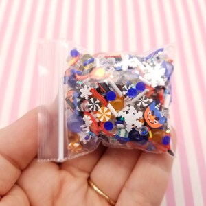 Peut inclure: Un sac en plastique transparent rempli de charmes en r&eacute;sine color&eacute;s. Les charmes sont en forme de flocons de neige, de cannes en sucre et d'autres motifs sur le th&egrave;me des f&ecirc;tes. Il y a aussi des paillettes bleues et orange.