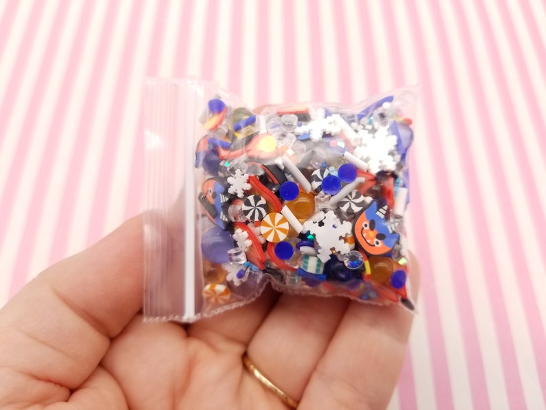 Peut inclure: Un sac en plastique transparent rempli de charmes en r&eacute;sine color&eacute;s. Les charmes sont en forme de flocons de neige, de cannes en sucre et d'autres motifs sur le th&egrave;me des f&ecirc;tes. Il y a aussi des paillettes bleues et orange.