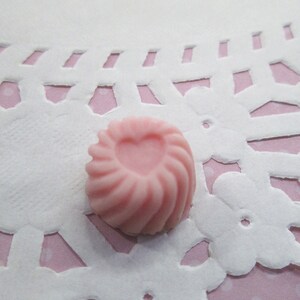 May include: A small, pink, swirl-shaped candy with a heart-shaped indentation in the center. The candy is set on a white, lace-like doily with a pink polka-dot background. The candy's texture appears smooth and the color is a soft pastel pink.