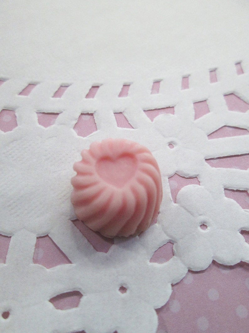 May include: A small, pink, swirl-shaped candy with a heart-shaped indentation in the center. The candy is set on a white, lace-like doily with a pink polka-dot background. The candy's texture appears smooth and the color is a soft pastel pink.