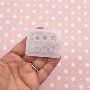 May include: A clear silicone mould in a rectangular shape, featuring star shapes of different sizes. The mould is held in a hand against a pink background with white polka dots. The stars are arranged in two rows.