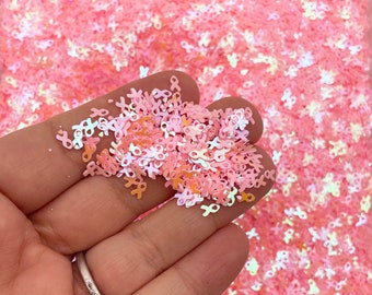 May include: Pink iridescent glitter in the shape of ribbons. The glitter is loose and scattered in a hand.