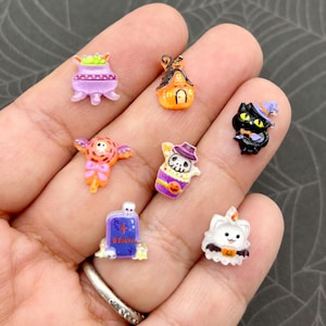 May include: A collection of miniature Halloween-themed charms. The charms include a purple cauldron, an orange haunted house, a black cat wearing a witch hat, a ghost, a tombstone, and a white cat with a pumpkin.