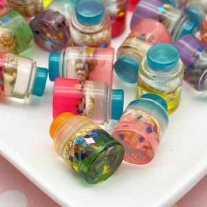 May include: A collection of small, colorful resin bottles with blue caps. The bottles contain decorative elements like seashells and glitter. The bottles are in shades of pink, green, yellow, and purple, and are arranged on a white surface.