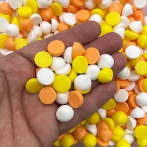 May include: A hand holding a variety of small, round, and cone-shaped candy decorations in white, yellow, and orange colors. The candy is scattered on a surface.