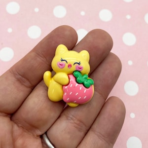 Könnte beinhalten: Eine gelbe Katzenfigur mit geschlossenen Augen und rosa Wangen, die eine rosa und grüne Erdbeere hält.