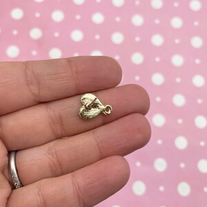 May include: A small, gold-colored heart-shaped charm with a loop for attaching to jewelry. The charm has a textured surface and a small post on the back. The charm is held in a person's hand against a pink background with white polka dots.