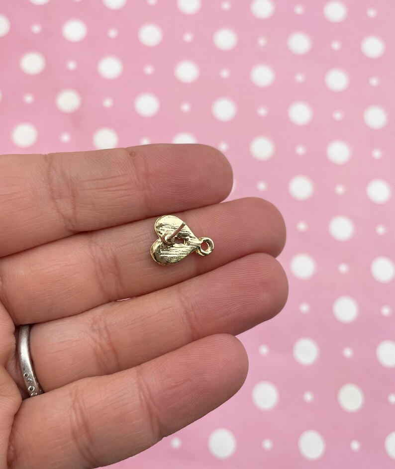 May include: A small, gold-colored heart-shaped charm with a loop for attaching to jewelry. The charm has a textured surface and a small post on the back. The charm is held in a person's hand against a pink background with white polka dots.