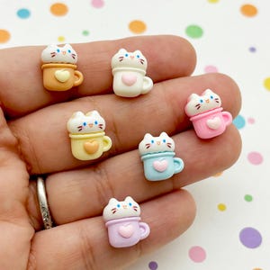 Op de afbeelding: Een verzameling van zeven miniatuur katvormige beeldjes in pastelkleuren. Elke kat zit bovenop een theekopje, met hartvormige details. De theekopjes zijn in tinten oranje, wit, geel, roze, blauw en lavendel.