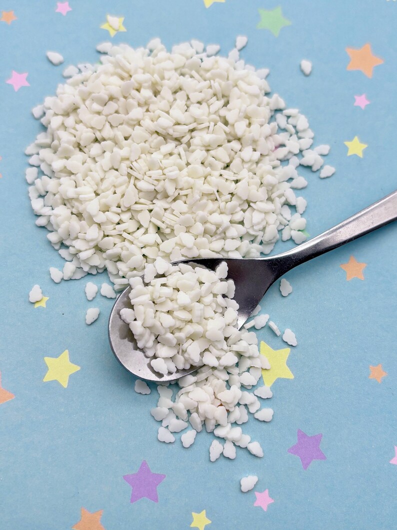 May include: A pile of small, white, irregularly shaped sprinkles next to a silver spoon. The sprinkles are on a light blue surface decorated with colorful star-shaped confetti.