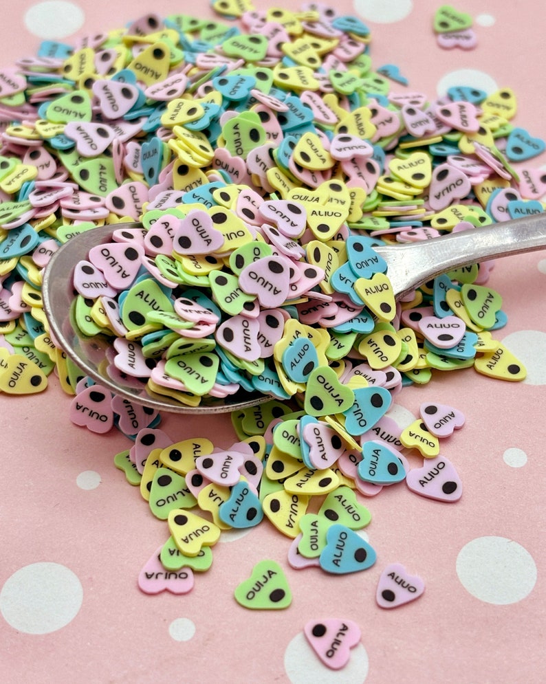 May include: A close-up of a spoon filled with colorful heart-shaped sprinkles. The sprinkles are decorated with black dots and the words "OUIJA" and "ALIUO".