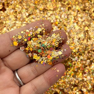 May include: A close-up of a hand holding a pile of gold glitter with iridescent and holographic finishes. The glitter is a mix of different sizes and shapes, creating a sparkling effect.