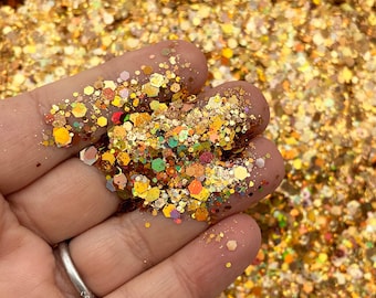 May include: A close-up of a hand holding a pile of gold glitter with iridescent and holographic finishes. The glitter is a mix of different sizes and shapes, creating a sparkling effect.