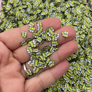 May include: A close-up of a hand holding a pile of small, yellow and silver, smiling face shaped resin embellishments. The embellishments are scattered on a surface covered in the same embellishments.