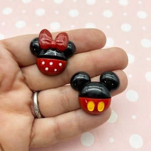 Puede incluir: Dos cabujones de resina de personajes de dibujos animados, uno es Mickey Mouse con orejas negras y una cara roja con círculos amarillos, el otro es Minnie Mouse con orejas negras y una cara roja con lunares blancos y un lazo rojo.