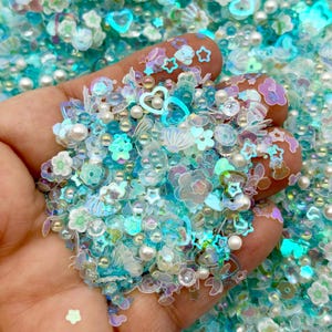 May include: A handful of iridescent sequins and beads in shades of blue, teal, and white. The mix includes flower, star, and heart shapes, along with small pearl beads. The sequins have a shimmering, translucent quality, creating a sparkling effect.