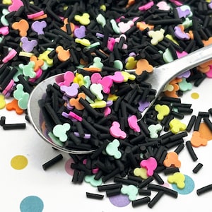 May include: A close-up of colorful sprinkles in a variety of shapes, including black sticks and small Mickey Mouse heads. The sprinkles are in a silver spoon and scattered on a white surface with colorful polka dots.