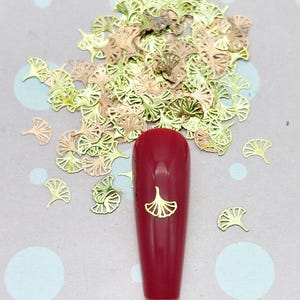 May include: A collection of small, metallic ginkgo leaf-shaped nail art charms in gold and rose gold tones, scattered around a single, long, glossy dark red artificial fingernail that features one matching gold ginkgo leaf charm applied to its center.