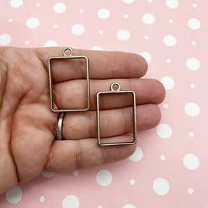 May include: Two silver-toned metal rectangular pendant frames with a loop at the top. The frames are different sizes.
