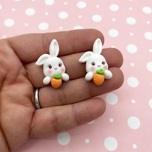 May include: Two white bunny charms holding orange carrots. The bunnies have pink cheeks and black eyes.