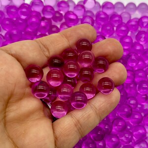 May include: A hand holding a handful of small, translucent purple water beads. The beads are round and smooth, and they are scattered across a surface.
