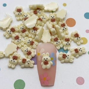 May include: A selection of small, off-white bunny-shaped nail art decorations with brown bows. Some are scattered, while others are on a pink nail with flower decals. The background is a white surface with colourful polka dots.